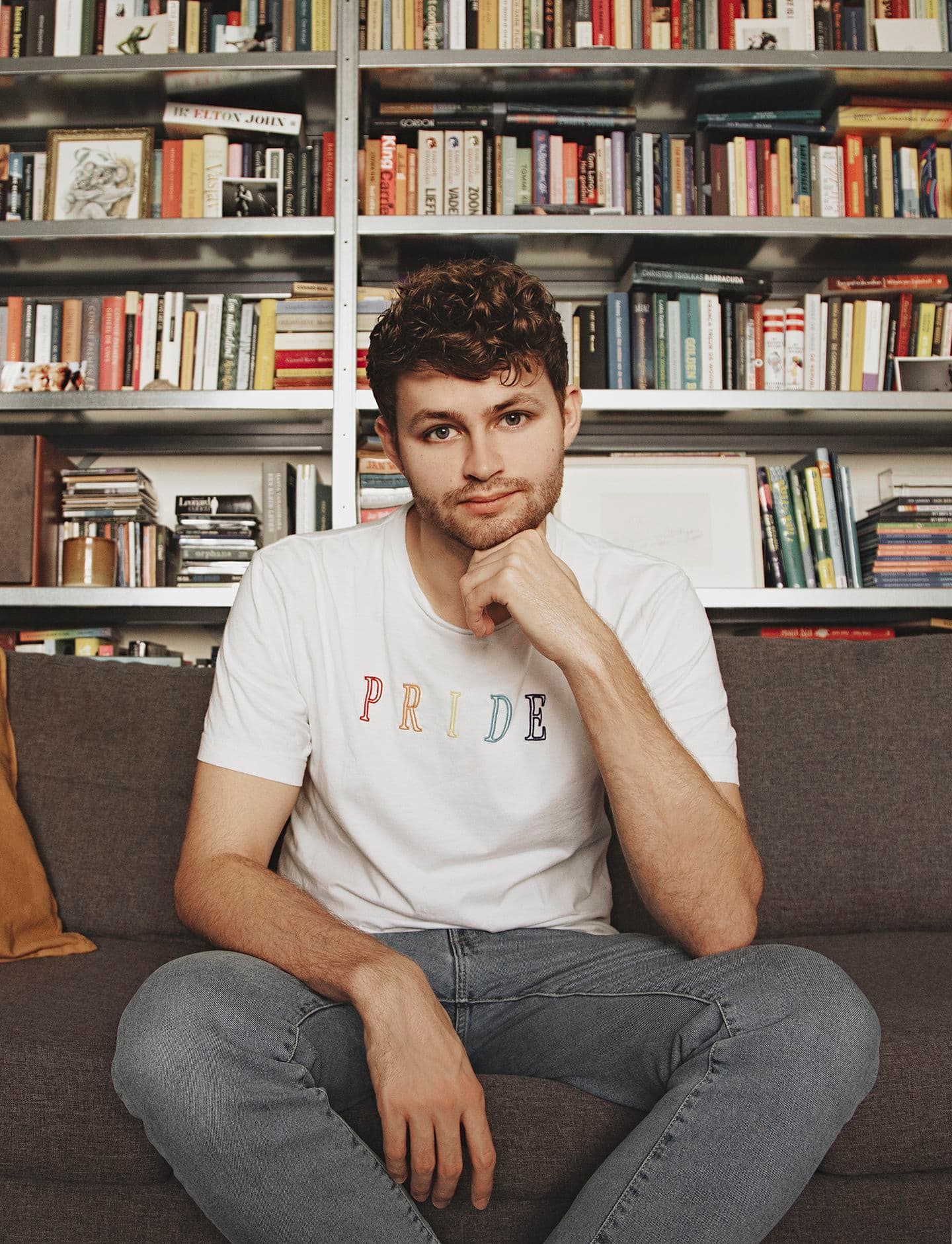 Pim Lammers zittend op de bank, voor zijn boekenkast. Hij leunt op zijn hand en heeft een T-shirt aan waar 'Pride' op staat in regenboogkleuren.